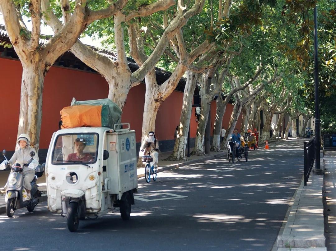 Shaded street beside De Shou Palace, with red walls, cyclists, and lush trees.