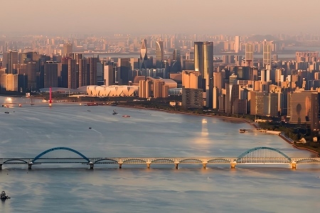 View of Qiantang River with arch railway bridge, dense modern skyscrapers, Hangzhou Olympic Sports Center and calm river surface under warm sunset glow