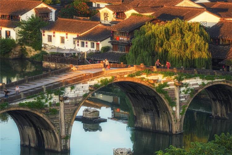 Three-arched ancient stone Gongchen Bridge crossing the Grand Canal, traditional white-washed old houses and weeping willows under warm sunset light in Hangzhou