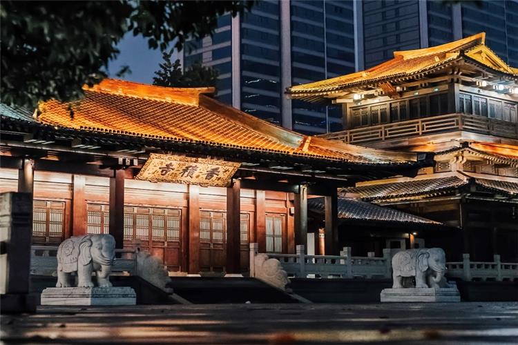 Lit ancient wooden halls of Xiangji Temple with stone elephant sculptures at the gate, classic Chinese Buddhist architecture against modern city buildings at dusk
