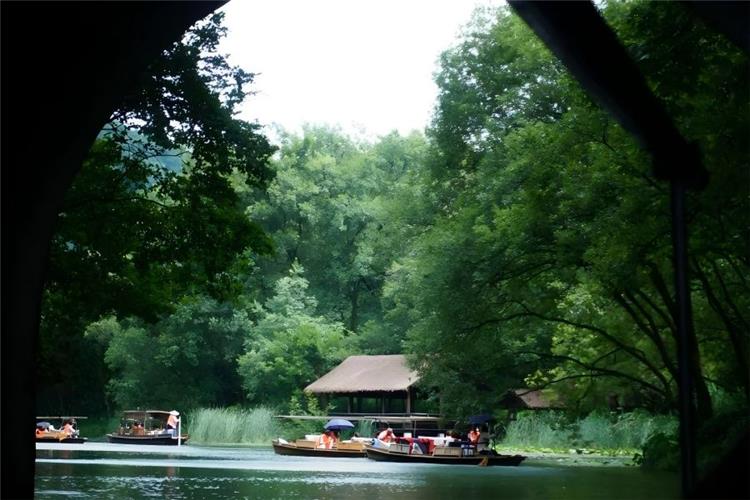 Traditional yuloh boats drifting on quiet Turtle Pool of West Lake Hangzhou, framed through ancient stone arch bridge tunnel, surrounded by lush green forest, thatched waterside pavilions and mirror-like emerald lake water
