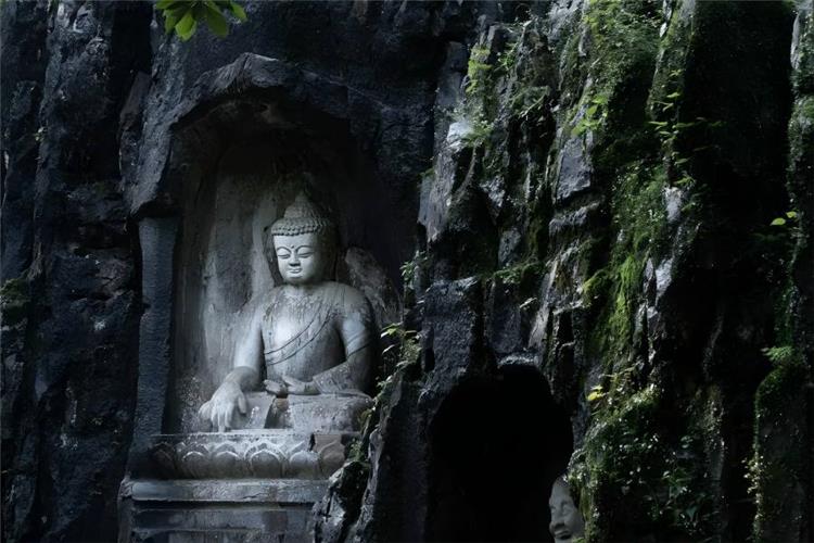 Weathered stone Buddha sculpture in Feilai Peak grotto, carved deep inside rugged dark cliff, green moss and wild plants covering rock walls, retaining primitive solemn charm of Southern Song Buddhist stone art