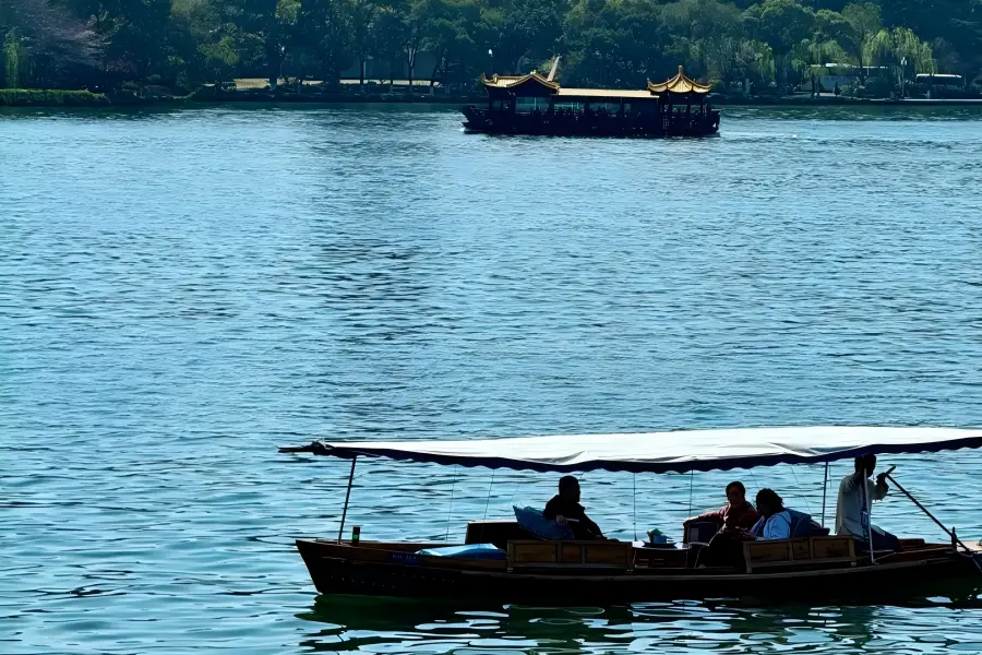 Sightseeing Boat to Three Pools Mirroring the Moon Elegant ancient-style West Lake pleasure boats depart from lakeside piers, cruising past islets and stone pagodas. Soft lake wind carries lotus fragrance, with layered green mountains forming a peaceful ink-wash backdrop all around.