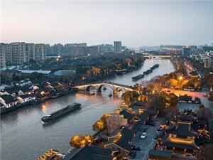 Stunning aerial dusk scenery of Gongchen Stone Arch Bridge along the ancient Grand Canal, featuring traditional Chinese architecture, river cargo boats and illuminated ancient streets in Hangzhou