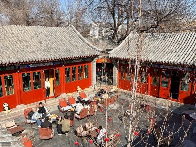 Classic red wooden Beijing siheyuan courtyard with tourists resting under clear sunny sky, ancient Chinese architecture