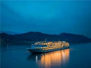 Illuminated luxury cruise vessel sailing on calm Yangtze River, dark mountain backdrop at blue hour dusk