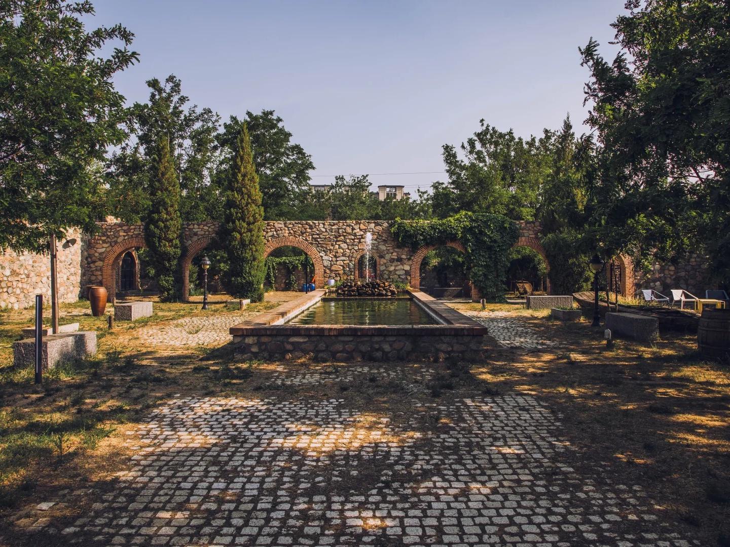 Sun?lit winery courtyard featuring central fountain, stone arch walls, cypress trees and cobblestone ground